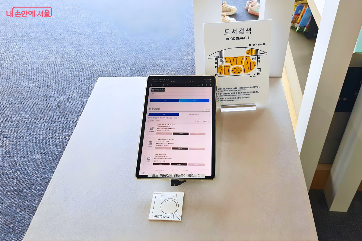 흰색 받침대 위에 놓인 태블릿 형태의 도서 검색 기기이다. 화면에는 도서 검색 결과 리스트가 나타나 있으며, 기기 아래에는 '들고 이동하면 경보음이 울립니다'라는 경고 문구가 붙어 있다. 기기 뒤쪽에는 도서관 평면도와 함께 주황색으로 검색 위치가 표시된 '도서검색 BOOK SEARCH' 안내판이 서 있다.