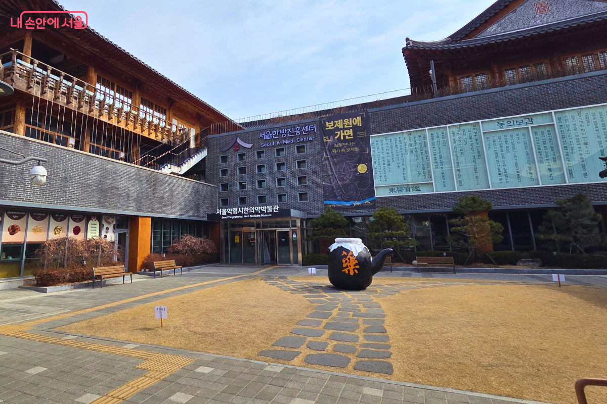 센터 건물들로 둘러싸인 야외 광장 마당의 모습이다. 마당 한가운데에는 '藥(약)'이라는 주황색 글자가 크게 새겨진 대형 검은색 약탕기 모양 조형물이 놓여 있으며, 바닥에는 돌판이 깔린 산책로가 나 있다. 정면에는 '서울약령시한의약박물관' 입구가 보이고, 건물 외벽 유리창에는 동의보감 관련 텍스트들이 빼곡하게 적혀 있다.