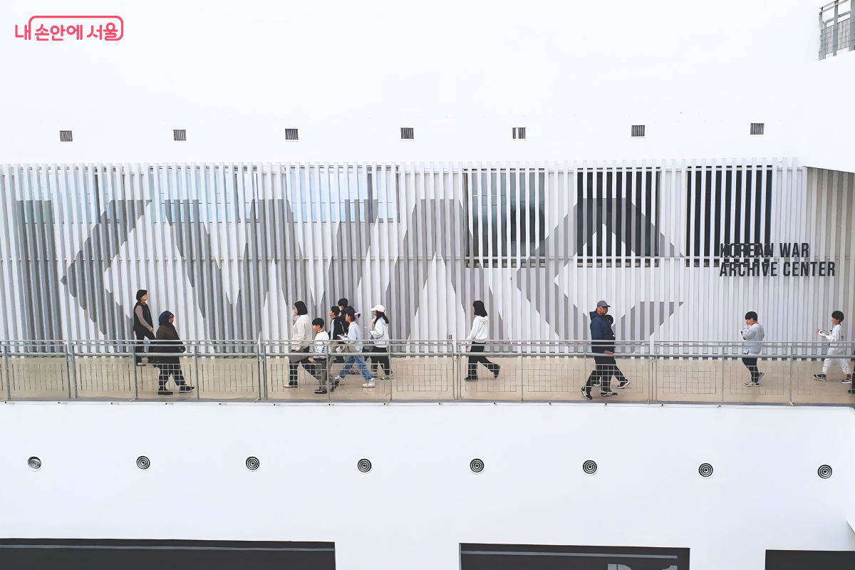 전쟁기념관 내부의 높은 층에서 아래를 내려다본 복도 전경이다. 흰색 바탕의 벽면에 얇은 수직 창살 형태의 구조물이 덧대어져 있으며, 그 뒤로 회색의 커다란 'KWAC' 글자와 'KOREAN WAR ARCHIVE CENTER' 문구가 적혀 있다. 난간이 설치된 복도를 따라 성인과 어린이들이 줄을 지어 라이브러리 방향으로 걸어가고 있다. 