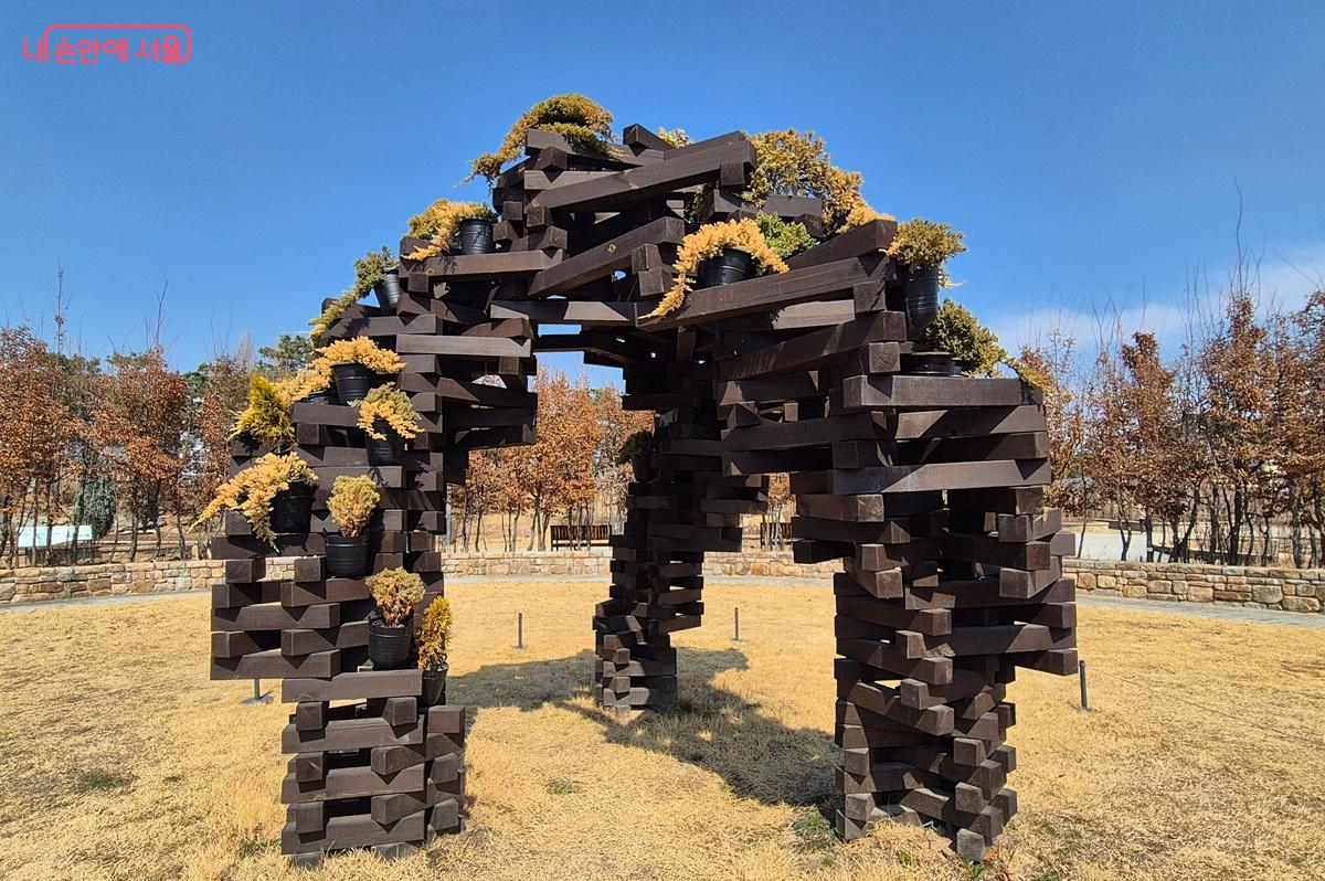 김주현 작가의 '생명의 그물-나무쌓기' 작품이 주제원에 전시되어 있다. 나무 토막을 쌓아 올려 아슬아슬 해보이면서도 견고한 구조에 식물들이 자라나고 있다. ⓒ마가은