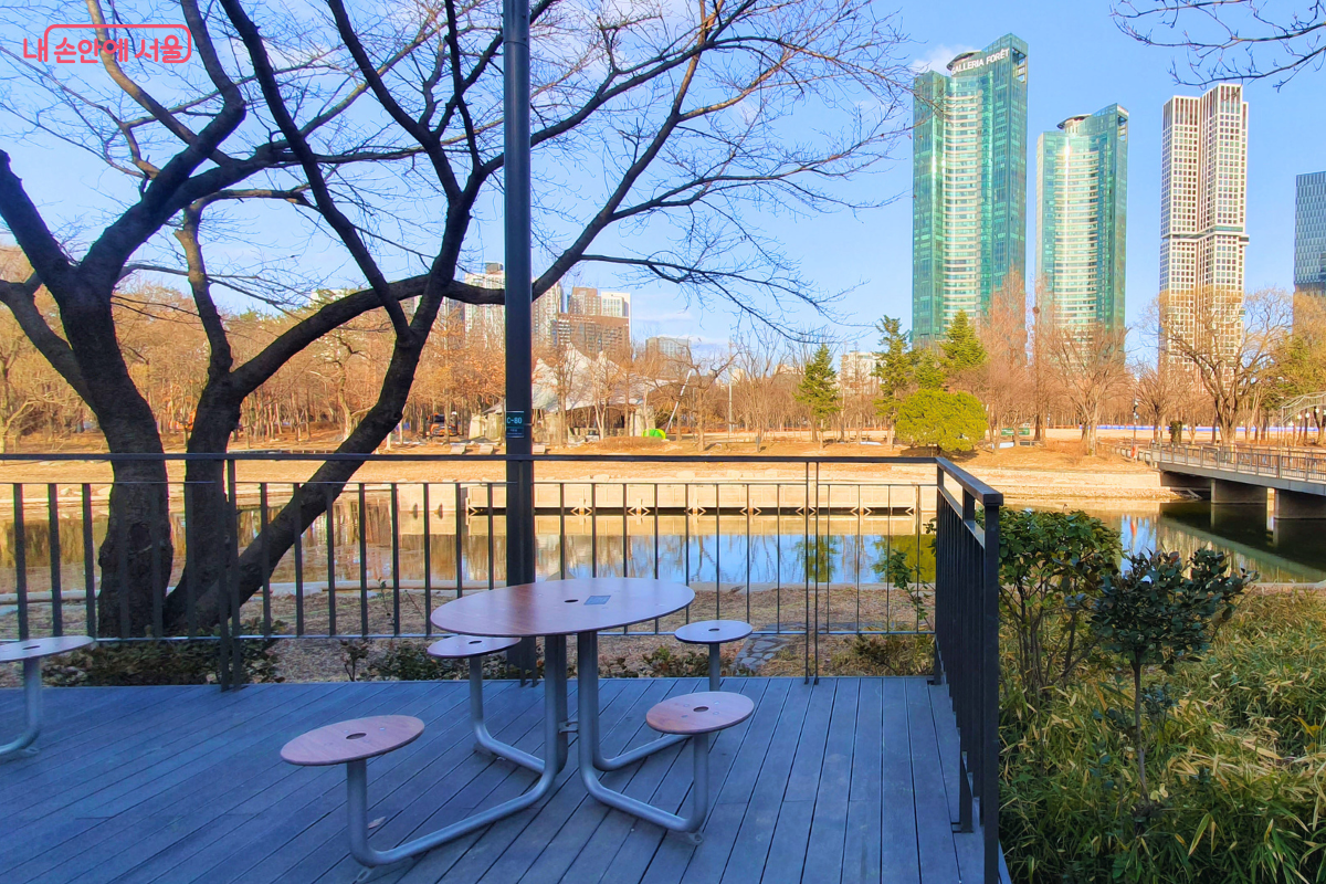 카페 앞 야외 테라스 좌석에서 바라본 풍경이다. 목재 데크 위에 타원형 야외 테이블과 의자가 배치되어 있으며, 그 너머로 잔잔한 서울숲 호수와 성수동의 랜드마크인 고층 빌딩들이 한눈에 들어온다. 호수 주변의 울창한 나무들과 현대적인 건축물이 어우러진 서울숲만의 독특한 수변 경관이 펼쳐져 있다. &copy;양정화