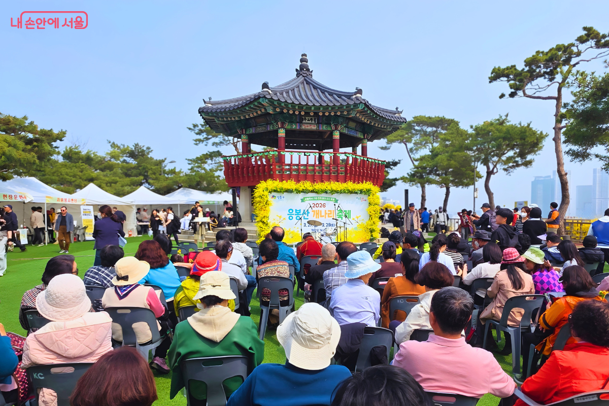 응봉산 정상 팔각정 앞에서 '2026 응봉산 개나리축제' 오프닝 행사가 열리고 있다. 팔각정 앞에는 개나리꽃으로 장식된 무대가 마련되어 있고, 수많은 시민이 의자에 앉아 무대를 지켜보고 있다. 주변에는 운영 부스들이 설치된 하얀 천막들이 보이며 푸른 잔디와 소나무가 어우러져 있다. &copy;신창근
