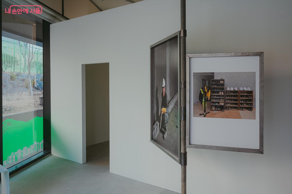 전시실 하얀 벽면에 공사 현장 작업자의 모습과 안전모, 작업화 등이 놓인 선반을 찍은 사진 작품 두 점이 나란히 전시되어 있다.