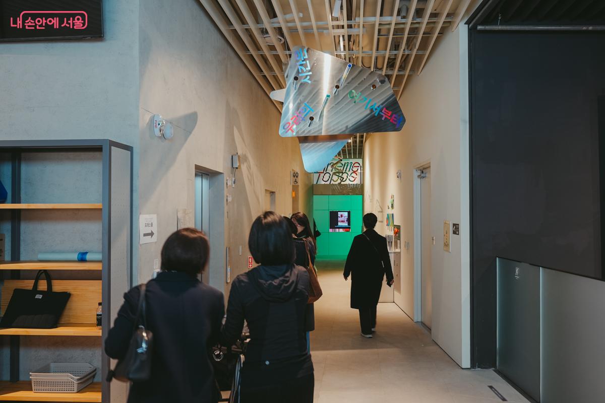 전시실 복도 천장에 '우리의 시간은 여기서부터'라는 다채로운 색상의 글씨가 적힌 은빛 모빌 간판이 매달려 있다.