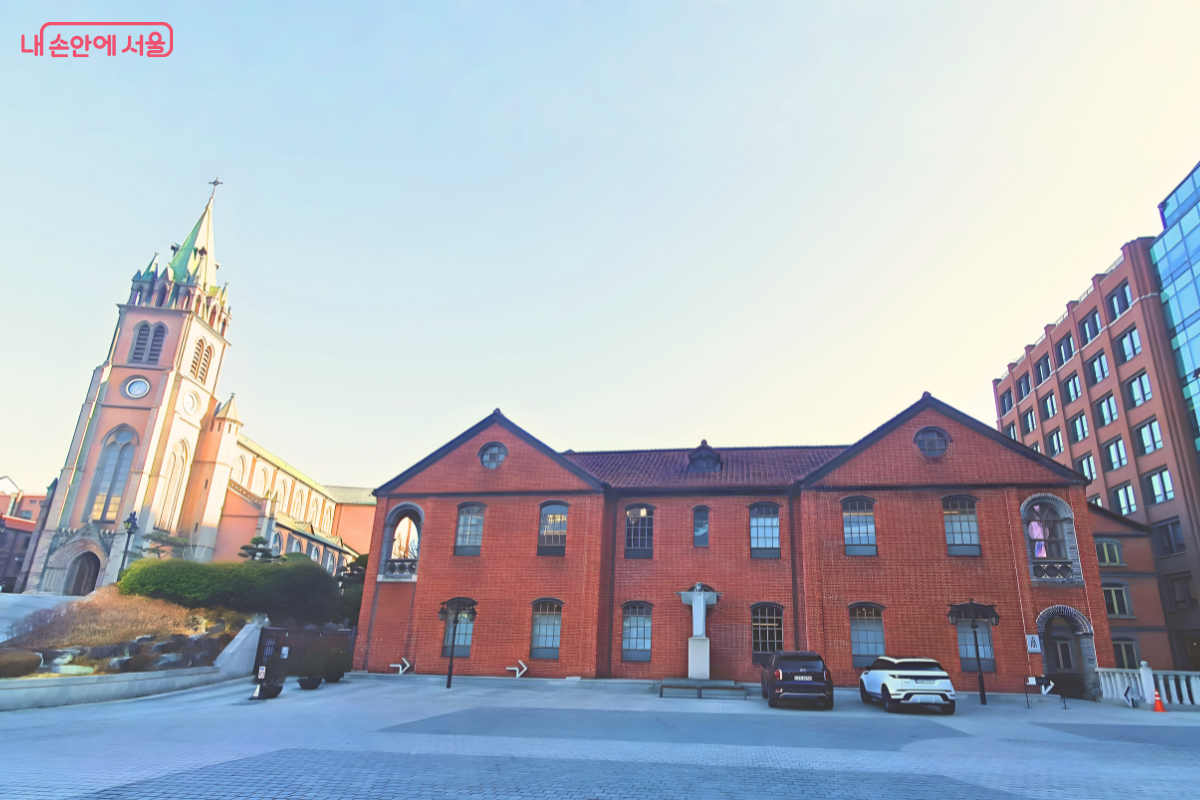 맑은 하늘 아래 명동대성당과 사도회관이 함께 보이는 넓은 구도의 사진이다. 왼쪽 언덕 위에는 명동대성당의 첨탑이 솟아 있고, 중앙에서 오른쪽으로는 2층 규모의 긴 붉은 벽돌 건물인 사도회관이 자리 잡고 있다. &copy;양정화