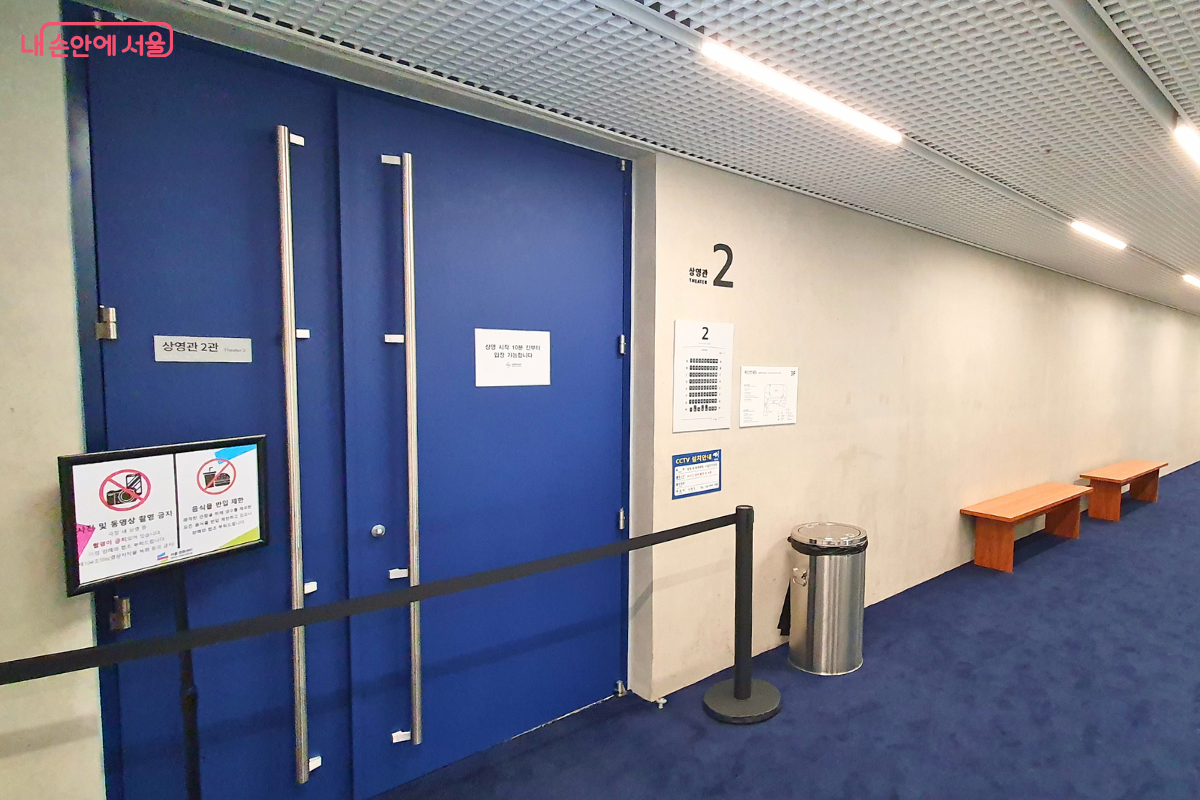 서울영화센터 내 상영관 2관의 입구 모습이다. 짙은 파란색의 커다란 이중문이 닫혀 있으며 문 옆벽에는 '상영관 2관'이라는 명칭과 함께 좌석 배치도가 부착되어 있다. 문 앞에는 사진 및 동영상 촬영 금지, 음식물 반입 제한 안내판이 세워져 있고 복도에는 방문객이 대기할 수 있는 나무 벤치가 놓여 있다. &copy;신창근