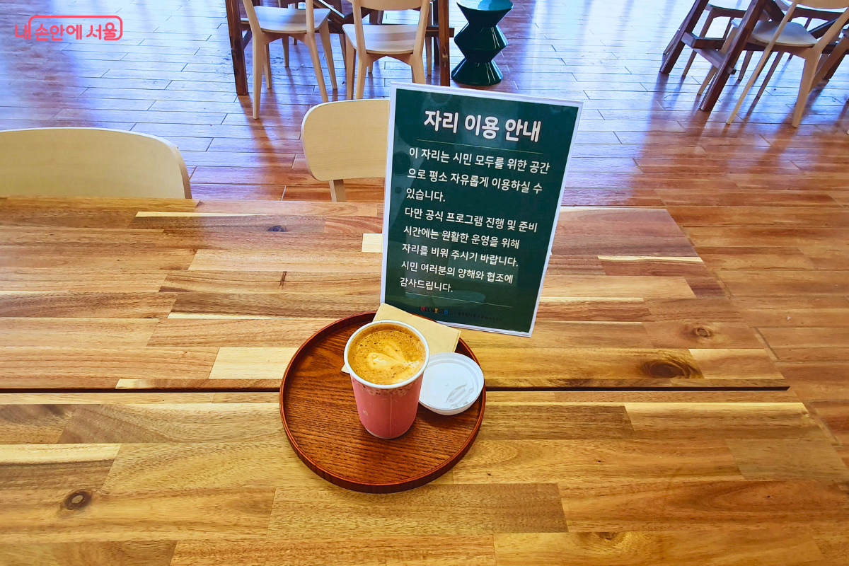 카페 내부 좌석의 모습이다. 원목 소재의 넓은 공용 테이블 위에 커피 한 잔과 &lsquo;자리 이용 안내&rsquo; 표지판이 놓여 있다. 안내문에는 이 자리가 시민 모두를 위한 자유로운 공간임을 밝히고, 공식 프로그램 진행 시 양해를 구하는 내용이 적혀 있다. 배경으로는 통유리창을 통해 들어오는 채광과 나무 의자들이 배치된 쾌적한 실내 전경이 보인다. &copy;양정화