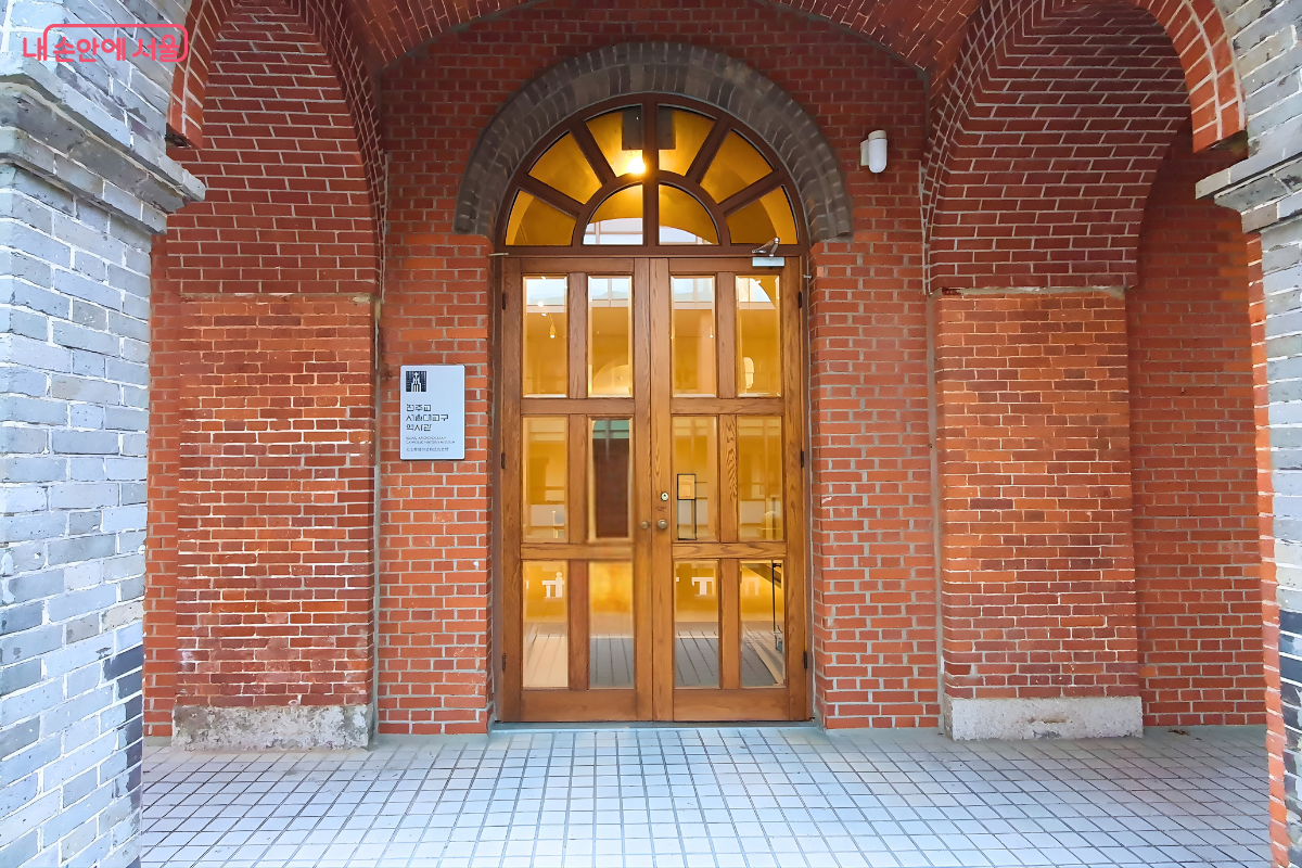 붉은 벽돌 아치 구조 안에 설치된 나무 재질의 주 출입구 정면 사진이다. 문 상단은 반원형 유리창으로 되어 있어 내부의 따뜻한 조명이 비치며, 문 왼쪽 벽면에는 '천주교 서울대교구 역사관 사도회관' 간판이 부착되어 있다. &copy;양정화