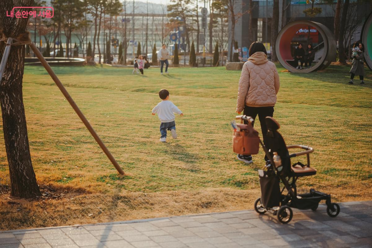 넓은 공원 잔디밭 위로 흰옷을 입은 어린아이가 신나게 뛰어가는 뒷모습이 보이고, 그 옆에는 유모차가 세워져 있다.