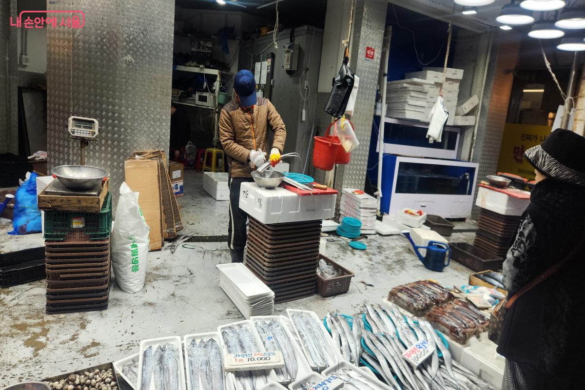 남구로시장 한 생선가게에서 상인이 갈치를 손질하고 있다. 작업대 위에 은빛 갈치가 놓여 있으며, 판매를 위해 손질 작업이 진행되고 있다.