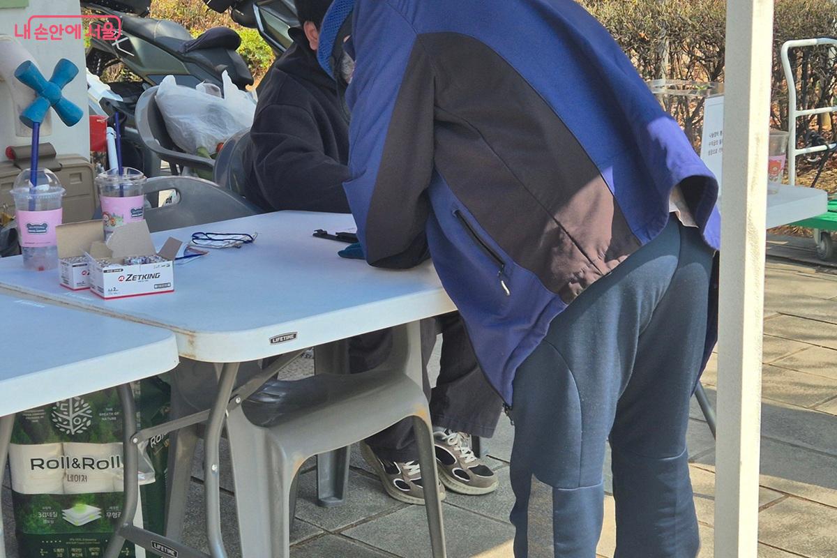 장터 입구에 마련된 안내데스크에서 한 어르신이 신청서를 작성하고 있다. 테이블 위에는 새 건전지가 있고, 테이블 아래로는 롤 휴지가 보인다. ©마가은
