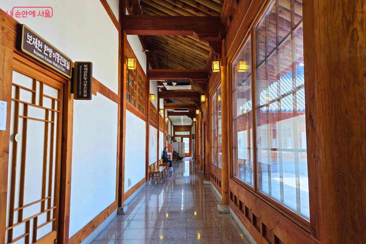 전통 한옥 양식의 서까래와 굵은 목재 보가 그대로 드러난 실내 복도다. 왼쪽 흰 벽에는 '보제원 한방이동진료실'이라고 적힌 갈색 명판이 부착되어 있고, 오른쪽은 통유리창을 통해 야외 마당이 보인다. 복도 중간에는 나무 벤치가 놓여 있으며, 천장에는 은은한 노란색 사각 한지 등이 일렬로 켜져 있다.