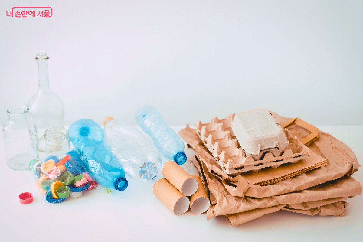 흰색 배경의 바닥에 종류별로 정갈하게 분류된 재활용 자원들이 놓여 있다. 왼쪽에는 내용물을 비운 투명한 유리병과 페트병들, 그리고 별도의 작은 비닐팩에 가득 담긴 알록달록한 플라스틱 병뚜껑들이 있다. 오른쪽에는 종이 재질의 자원들이 모여 있는데, 다 쓴 휴지 심 3개와 계란 판 1개, 그리고 여러 장의 갈색 종이 쇼핑백이 차곡차곡 쌓여 있다. 모든 자원은 이물질 없이 깨끗하게 세척된 상태로 분리되어 있다. 
