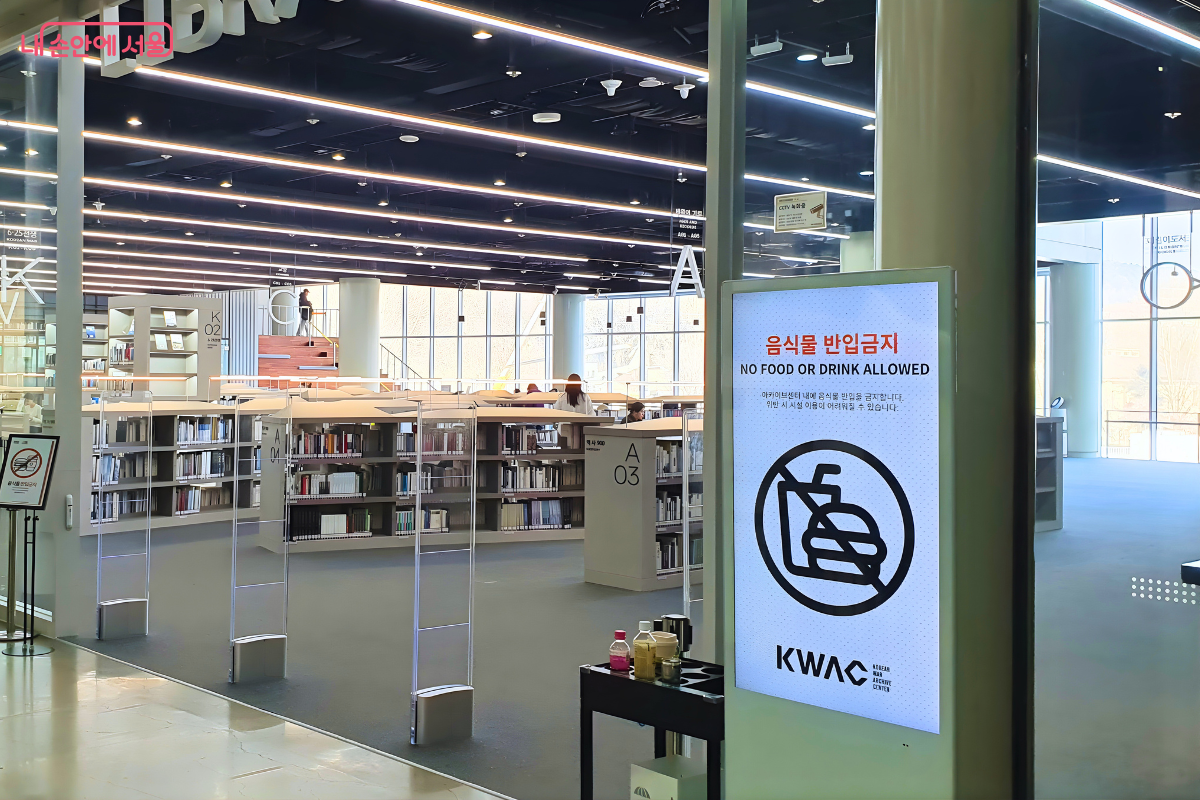 도서관 입구의 유리 벽과 입구 주변 모습이다. 우측 전면에는 '음식물 반입금지 NO FOOD OR DRINK ALLOWED'라는 문구와 아이콘이 그려진 세로형 안내 모니터가 서 있다. 유리문 너머로 도서관 내부의 낮은 서가들과 천장의 긴 조명들, 그리고 창가 쪽의 계단식 구조물이 보인다. 입구 바닥에는 금속 재질의 보안 게이트가 설치되어 있다.