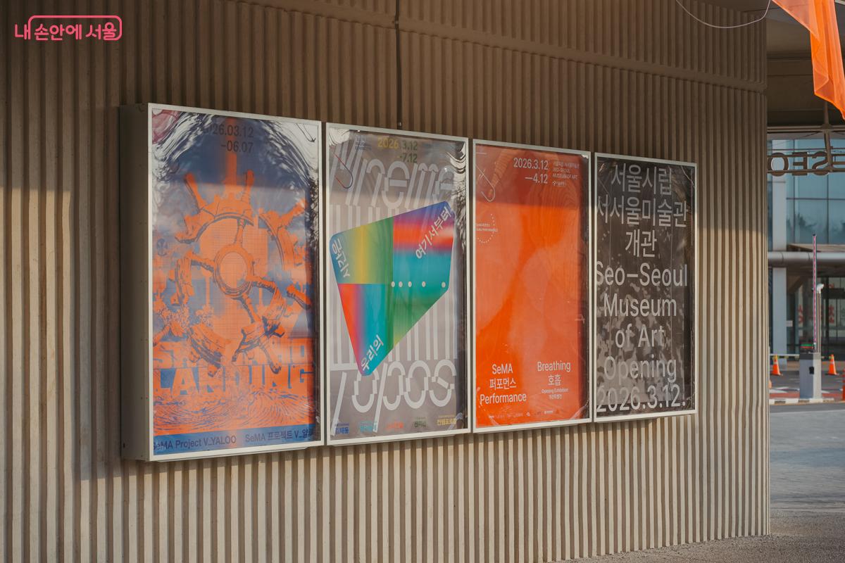 서서울미술관의 현대적인 회색 노출 콘크리트 외벽 코너에 부착된 개관 기념 건립기록전의 대형 홍보 포스터 전경. 포스터 상단에는 알록달록한 기하학적 도형들과 함께 '우리의 시간은 여기서부터'라는 제목이 크고 선명하게 적혀 있고, 그 아래로 'SeMA 서서울미술관 건립기록전'이라는 전시 명칭이 보인다.