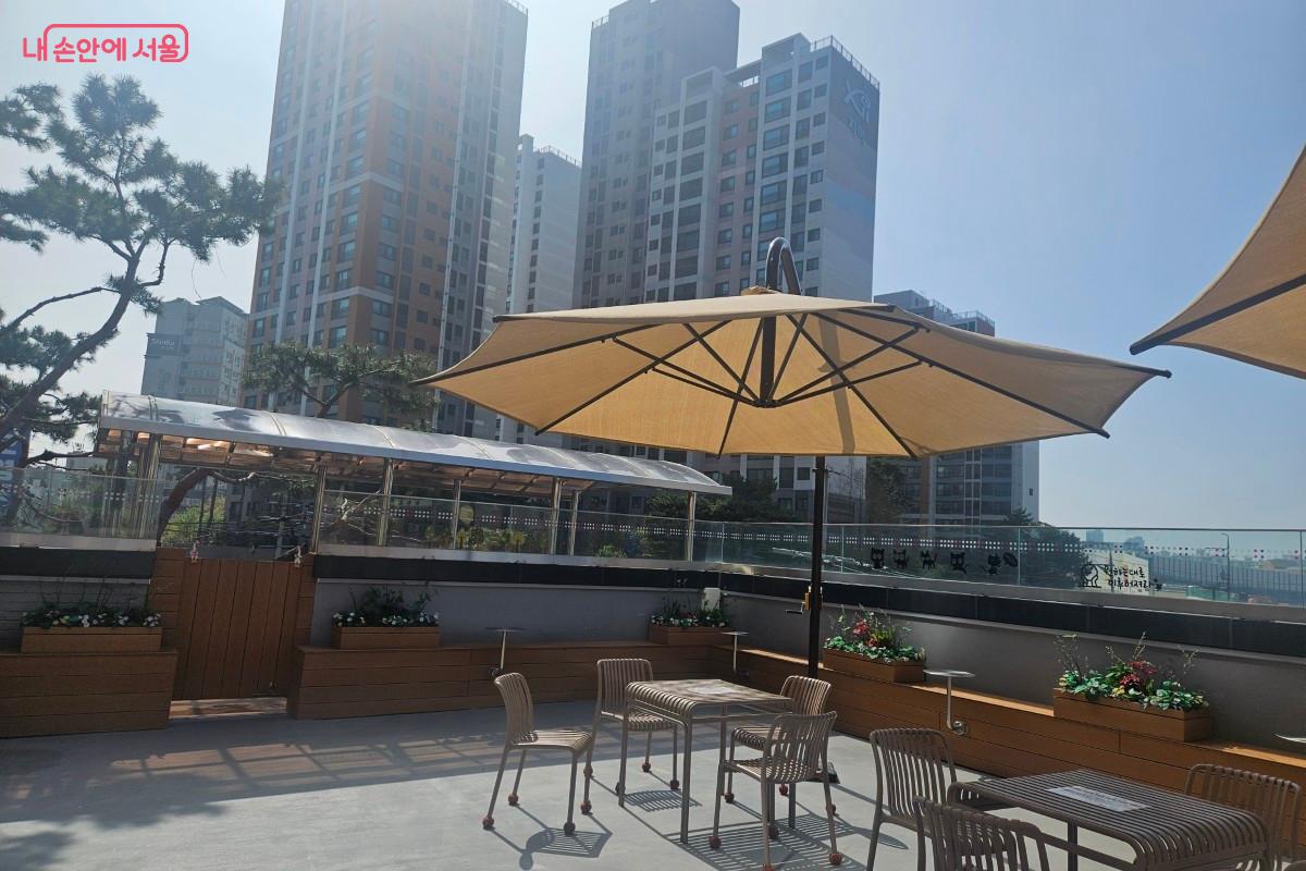 3층 야외테라스 전경, 뒤에는 아파트 전경이다. 파라솔이 햇빛을 가려주고 있으며, 4인이 앉을 수 있는 테이블이 적지 않게 자리잡고 있다.
