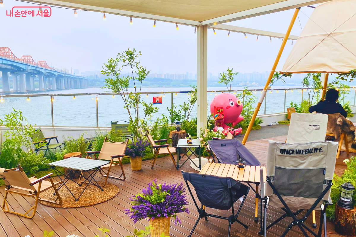 나무 데크가 깔린 루프탑의 모습이다. 다양한 형태의 캠핑 의자와 작은 야외용 테이블들이 여유롭게 배치되어 있다. 곳곳에 보라색 라벤더 화분과 초록색 식물들이 놓여 있으며, 강 쪽 유리 난간 앞에는 대형 분홍색 문어 캐릭터 인형이 설치되어 캠핑장 같은 분위기를 자아낸다.