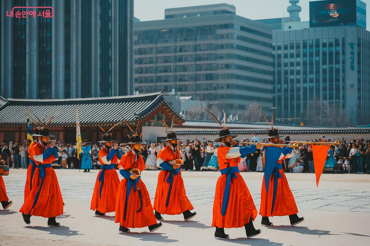 현대식 고층 건물들을 배경으로, 붉은 의상과 깃털 달린 모자를 쓰고 긴 나발을 불며 걸어가는 취타대원들 사진