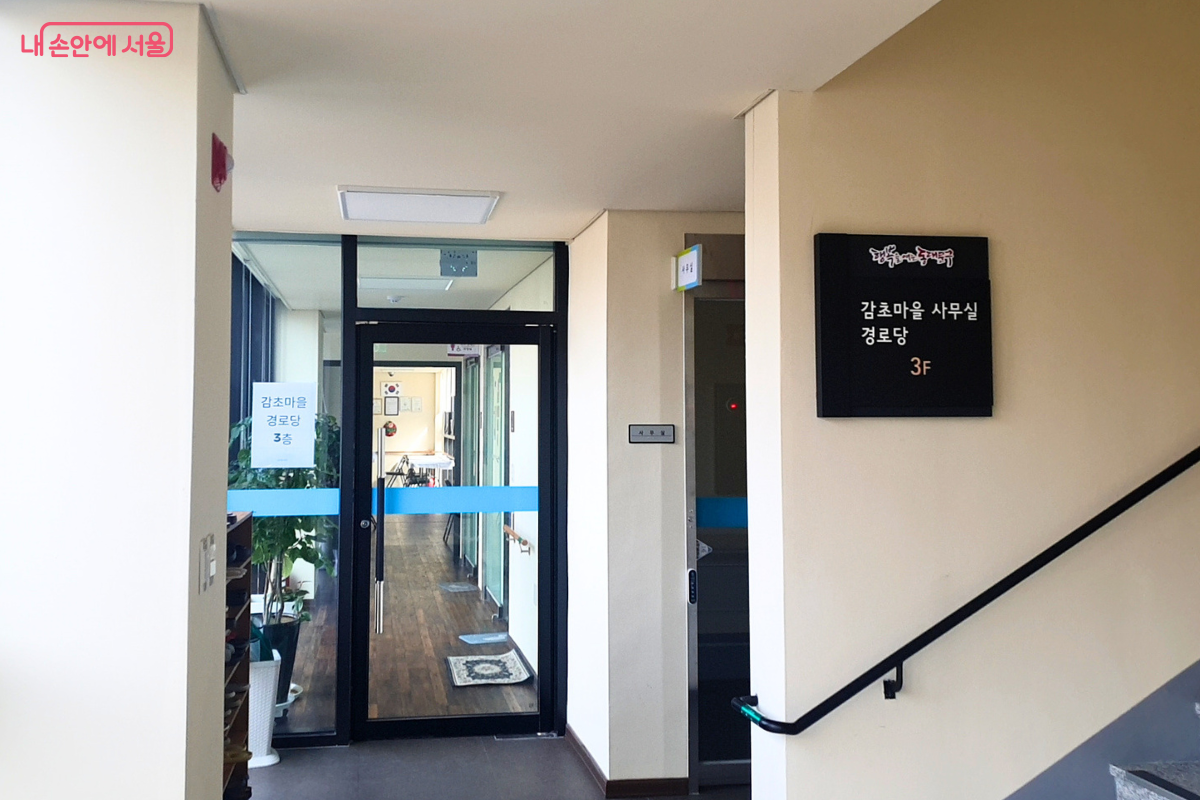 도서관 건물 3층의 입구 복도 전경이다. 왼쪽 유리문에는 '감초마을 경로당 3층'이라는 안내문이 부착되어 있고, 오른쪽 벽면에는 검은색 바탕에 하얀 글씨로 '감초마을 사무실 경로당 3F'라고 적힌 층별 표지판이 걸려 있다. 복도 끝으로는 경로당 내부의 밝은 조명이 보이며, 우측으로는 위층으로 올라가는 계단 손잡이가 설치되어 있다. 이곳은 지역 어르신들의 여가와 마을 행정 지원이 이루어지는 공간이다.
