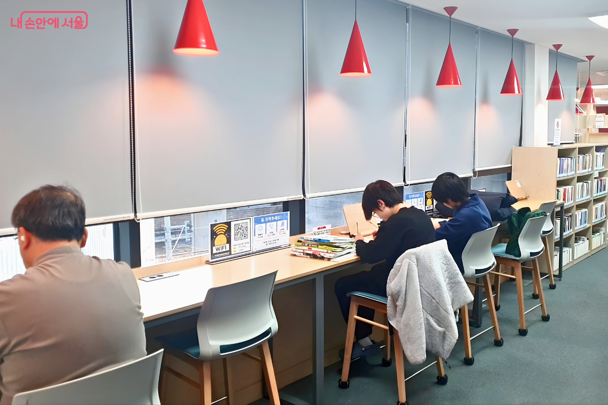 4층 제1열람실 창가에 마련된 1인용 데스크 자리의 모습이다. 외부 풍경을 마주하고 긴 테이블이 배치되어 있으며, 좌석마다 붉은색 고깔 모양의 펜던트 조명이 설치되어 집중도를 높여준다. 이용객들이 각자의 책상에서 독서와 학습에 몰입하고 있으며, 테이블 위에는 무료 와이파이 이용 안내문이 부착되어 있다.