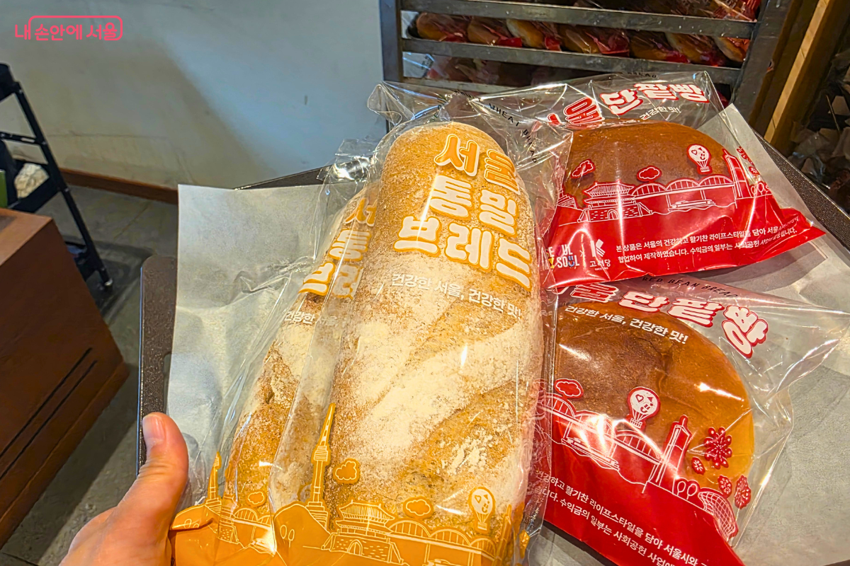 구매자가 빵 트레이 위에 직접 구매한 서울빵 4봉지를 담아 들고 있는 모습이다. 노란색 패키지의 '서울 통밀브레드' 2개와 붉은색 패키지의 '서울 단팥빵' 2개가 트레이 위에 놓여 있다. 패키지 전면에는 서울 시내의 스카이라인을 형상화한 일러스트와 제품명이 선명하게 인쇄되어 있다. 배경으로는 베이커리 매장 내부의 빵 진열 랙 일부가 보이며, 기자가 직접 구매를 인증하는 장면을 담고 있다.