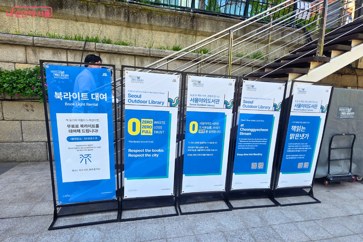 다섯 개의 안내판이 나란히 세워져 있다. 왼쪽부터 '북라이트 대여', 'Seoul Outdoor Library', '서울야외도서관', 'Cheonggyecheon Stream', '책읽는 맑은냇가' 안내판 순이다. 각 안내판은 파란색과 흰색 배경으로 구성되어 있다.