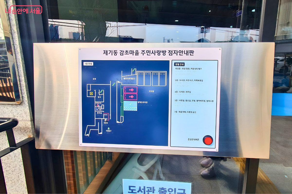 건물 1층 주출입구 벽면에 부착된 '제기동 감초마을 주민사랑방 점자 안내판'이다. 왼쪽에는 1층의 각 실 배치를 점자와 함께 입체적으로 표현한 지도가 그려져 있고, 오른쪽에는 옥상 정원부터 1층 주차장까지 각 층별 시설 명칭이 상세히 적혀 있다. 하단 우측에는 시각장애인을 위한 음성 안내를 시작하는 빨간색 버튼이 돌출되어 있어 무장애 환경을 구현하고 있다.