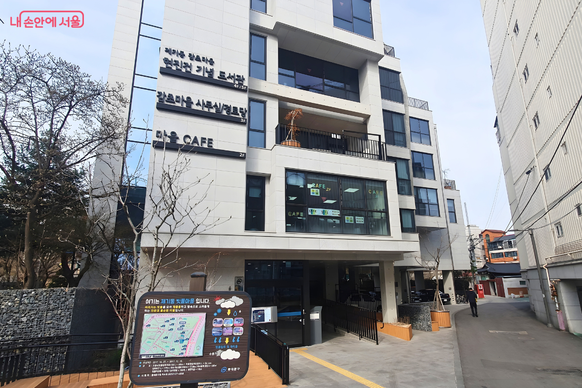 제기동 감초마을 현진건 기념 도서관 건물의 전체적인 외관 사진이다. 현대적인 백색 외장재로 마감된 지상 5층 규모의 건물로, 건물 외벽에는 각 층의 용도를 알리는 표지판이 부착되어 있다. 4층과 5층은 도서관, 3층은 사무실과 경로당, 2층은 마을 카페로 사용됨을 알 수 있다. 건물 앞에는 빗물을 재활용하는 '제기동 빗물마을' 안내판이 세워져 있다.