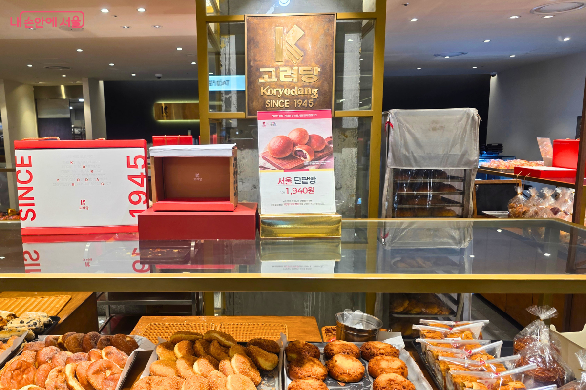 고려당 매장 내부의 전경으로, 기둥 상단에 'KORYODANG SINCE 1945'라고 적힌 빈티지한 스타일의 브랜드 현판이 부착되어 있다. 현판 아래에는 서울 단팥빵 1개의 가격이 1,940원임을 알리는 포스터가 부착되어 있으며, 진열장 위에는 'SINCE 1945'와 'KORYODANG' 로고가 새겨진 붉은색과 흰색 구성의 전용 선물 박스들이 쌓여 있다. 매대 전면에는 다양한 종류의 갓 구운 빵들이 바구니에 담겨 진열되어 있다.