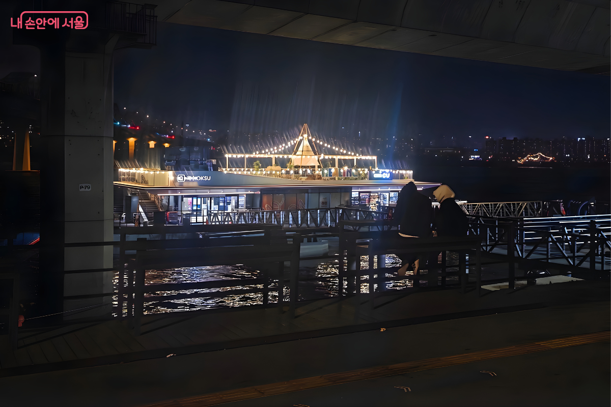한강 공원 둔치에서 바라본 옥수선착장의 모습이다. 어두운 배경 속에서 선착장 루프탑의 노란 조명이 유독 밝게 빛나고 있다. 화면 앞쪽 어두운 벤치에는 두 명의 시민이 앉아 강 너머 환하게 불을 밝힌 선착장 건물을 바라보고 있는 뒷모습이 담겨 있다.