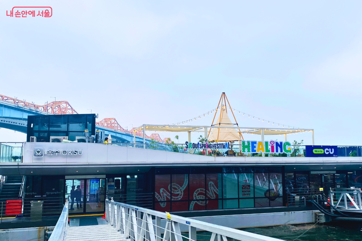 옥수선착장 건물의 측면 모습이다. 건물 왼쪽 상단에는 '옥수 OKSU'라는 간판이 붙어 있고, 루프탑 위에는 하얀색 캐노피와 원뿔 모양의 텐트가 설치되어 있다. 텐트 주변으로는 노란색 알전구들이 줄지어 장식되어 있으며, 건물 아래층은 유리 통창으로 되어 있다.