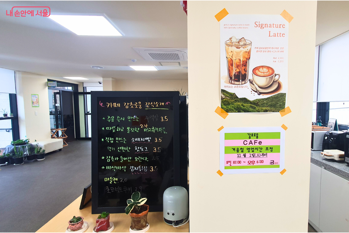 카페 한편에 마련된 간식 메뉴판과 영업시간 안내 포스터들이다. 검은색 칠판 메뉴판에는 '카페 감초로움 간식소개'라는 문구와 함께 주문 즉시 만드는 소시지빵(3.5), 핫도그(3.5), 감초가 들어간 오란다(1.5), 바삭바삭 감자튀김(3.5) 등의 저렴한 가격표가 적혀 있다. 우측 벽면에는 'Signature Latte' 홍보 포스터와 함께 11월 1일부터 적용되는 겨울철 영업시간(오전 10:00 ~ 오후 6:00, 금요일 휴무) 안내문이 부착되어 있다.