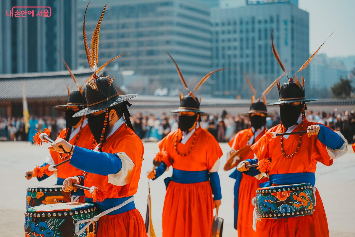 검은 천으로 얼굴을 가리고 붉은 옷을 입은 수문군들이 각을 맞춰 작은 북을 치고 있는 클로즈업 사진