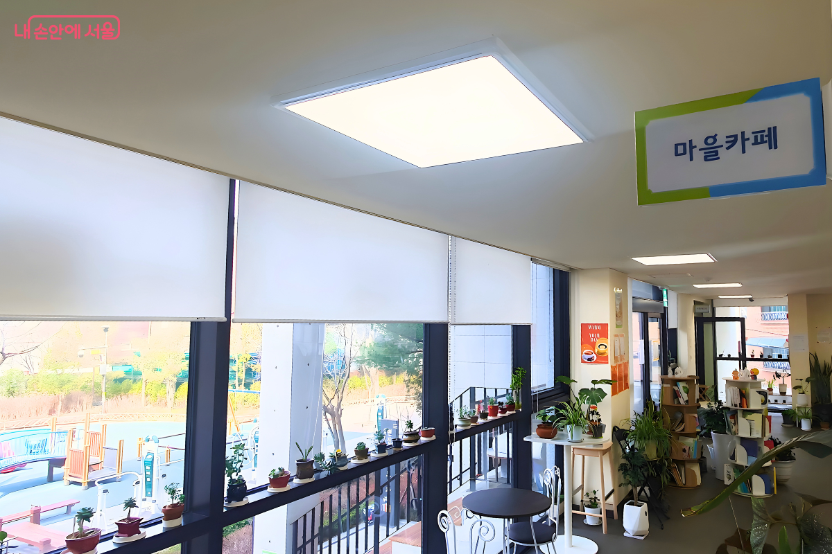도서관 건물 2층에 위치한 마을 카페 내부의 창가 쪽 풍경이다. 넓은 통창을 통해 밝은 햇살이 들어오고 있으며, 창틀 선반 위에는 작고 귀여운 다육식물 화분들이 나란히 놓여 있다. 천장에는 '마을카페'라고 적힌 연두색 테두리의 안내판이 매달려 있고, 창밖으로는 인접한 어린이 공원의 놀이기구 시설이 내려다보여 평화로운 마을 분위기를 느낄 수 있다.