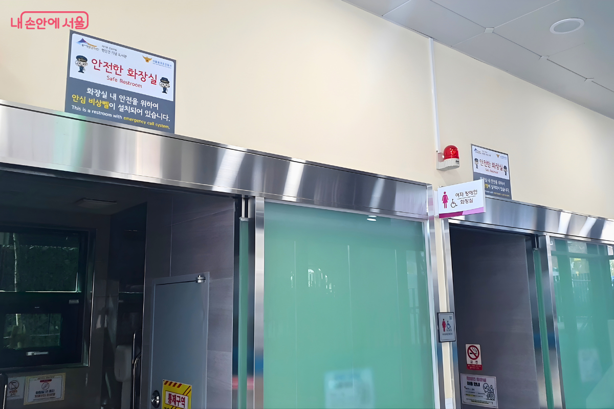 도서관 건물 1층에 설치된 '안전한 화장실' 입구 모습이다. 스테인리스 재질의 문 위로 '안전한 화장실(Safe Restroom)' 안내판이 부착되어 있으며, 화장실 내 안전을 위한 안심 비상벨 설치 문구와 음성 안내 시스템 정보가 적혀 있다. 문 옆으로는 시각장애인을 위한 성별 구분 안내와 함께 '여자 장애인 화장실'임을 알리는 표지판과 빨간색 안심 벨이 설치되어 있어 사용자 안전을 위한 세심한 설계를 보여준다.