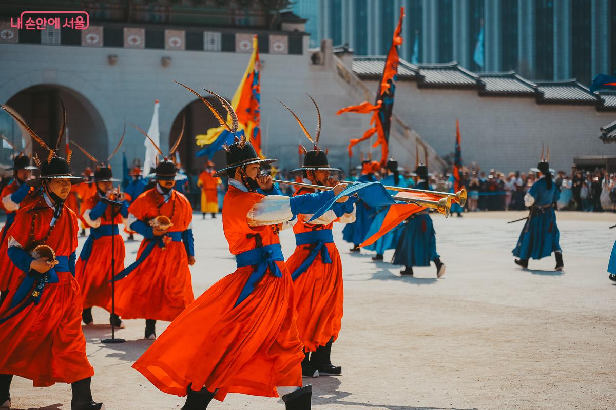 바람에 붉은 옷자락이 휘날리는 가운데 두 손으로 나발을 힘차게 불고 있는 대원의 역동적인 클로즈업 사진