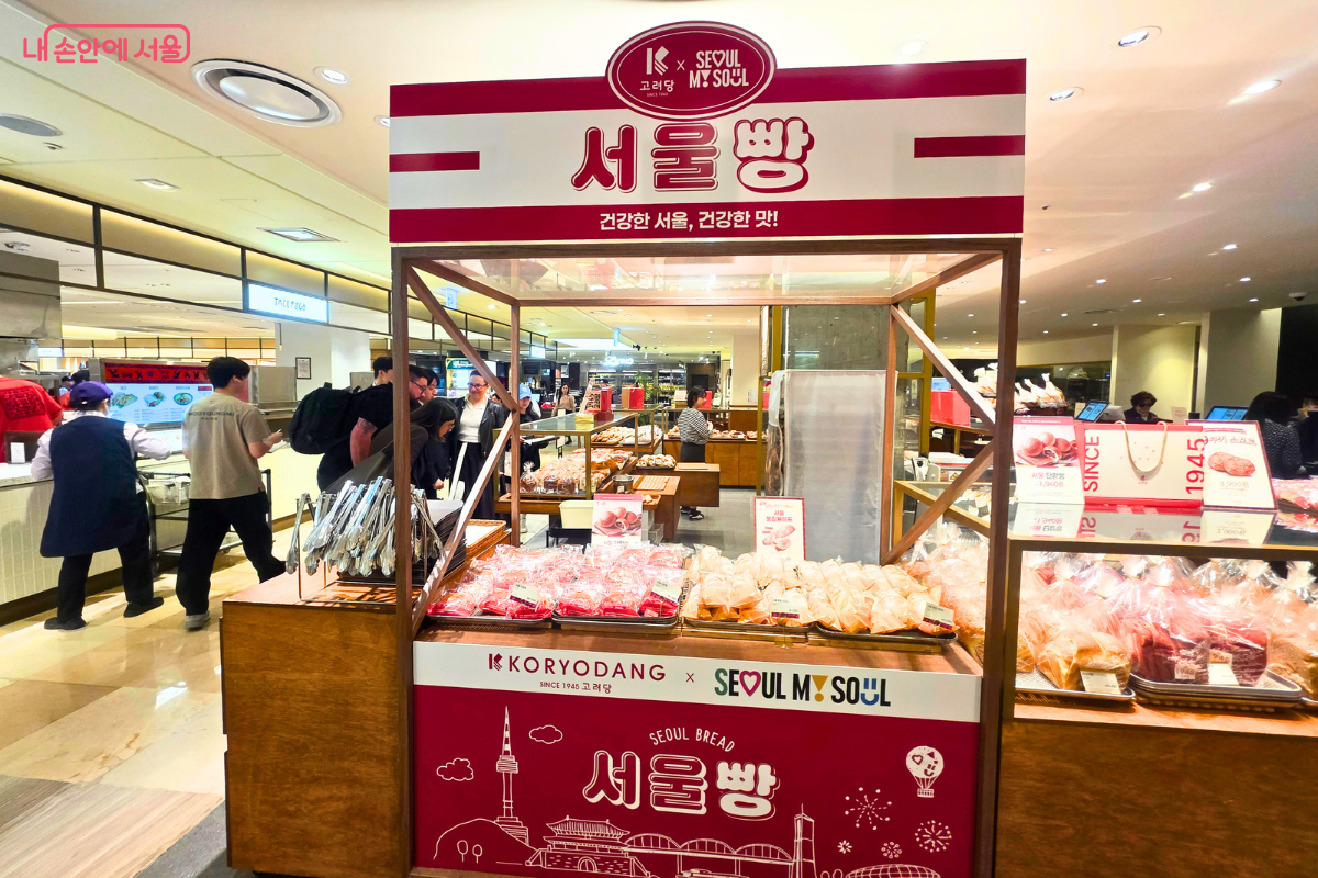 롯데백화점 본점 지하 1층에 위치한 고려당 매장 내 '서울빵' 전용 팝업 매대의 전경이다. 매대 상단에는 'KORYODANG x SEOUL MY SOUL' 로고와 함께 큰 글씨로 '서울빵'이 적혀 있으며, 그 아래로 '건강한 서울, 건강한 맛!'이라는 슬로건이 보인다. 매대 하단 전면부에는 붉은색 바탕에 남산서울타워, 숭례문, 롯데월드타워 등 서울의 주요 랜드마크 일러스트가 화이트 라인으로 그려져 있다. 매대 선반 위에는 투명한 비닐에 개별 포장된 빵들이 가득 진열되어 있다.