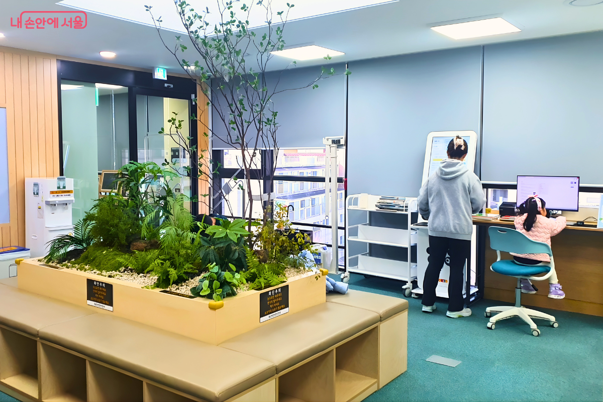 제기동 감초마을 현진건 기념 도서관 5층에 위치한 제2열람실 내부 전경이다. 열람실 중앙에는 초록색 식물들이 심어진 실내 화단이 조성되어 있어 쾌적한 분위기를 자아내며, 화단 주변은 앉아서 쉴 수 있는 벤치 형태의 공간으로 구성되어 있다. 한쪽 공간에서는 어린이 이용객이 컴퓨터를 사용하고 있으며, 자가 대출 반납기 등 최신 도서 이용 장비가 구비되어 있다.