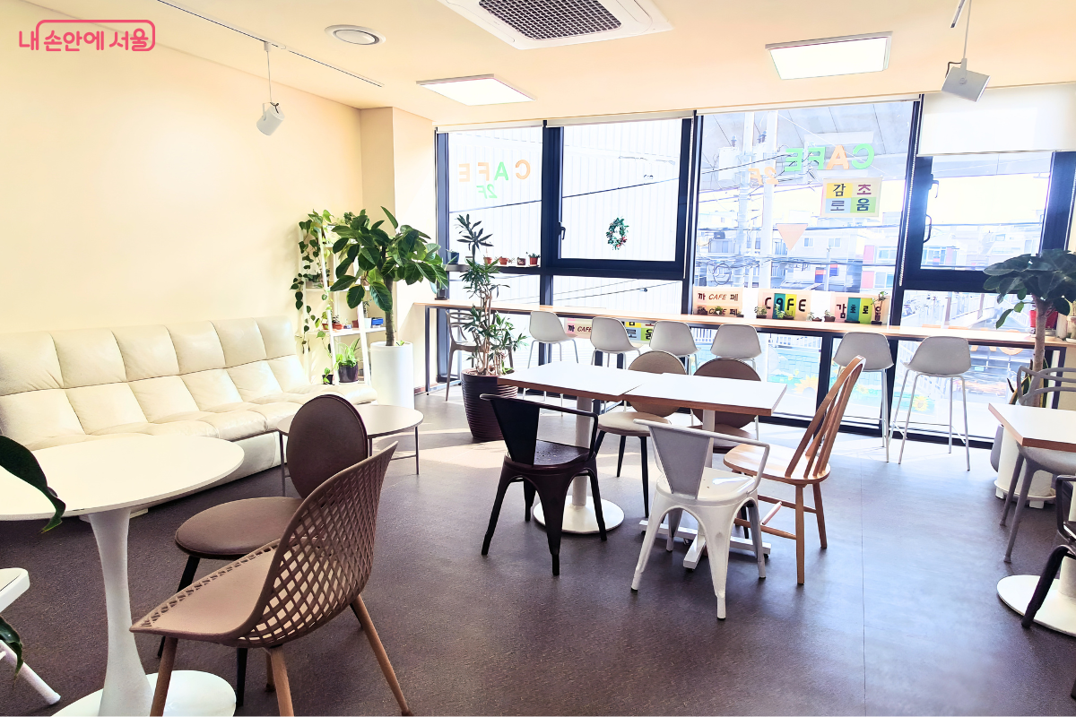 카페 내부의 전체적인 좌석 배치 모습이다. 한쪽 벽면에는 푹신한 하얀색 가죽 소파가 놓여 있고, 중앙에는 원형 및 사각형의 하얀색 테이블과 모던한 디자인의 의자들이 여유 있게 배치되어 있다. 창가 쪽은 바(Bar) 형태의 긴 테이블과 높은 의자들이 마련되어 공부나 독서를 하기에 적합해 보이며, 유리창에는 'CAFE 감초로움'이라는 카페 이름이 알록달록한 글자로 부착되어 있다.