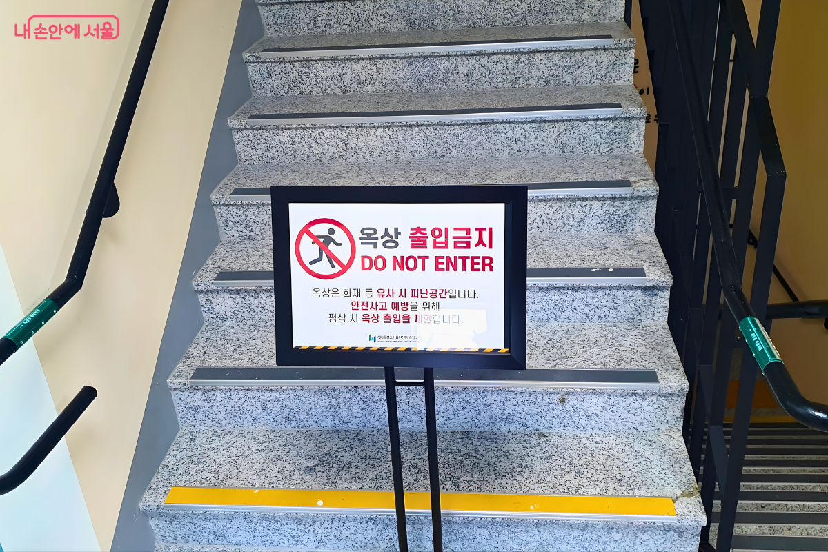 옥상으로 올라가는 계단 입구에 설치된 출입 통제 안내판이다. 검은색 프레임의 거치대 안에는 빨간색 금지 기호와 함께 '옥상 출입금지(DO NOT ENTER)'라는 문구가 크게 적혀 있다. 안내판 하단에는 '옥상은 화재 등 유사시 피난 공간이며, 안전사고 예방을 위해 평상시 옥상 출입을 제한한다'는 설명이 명시되어 있어 도서관의 철저한 안전 관리 수칙을 보여준다.
