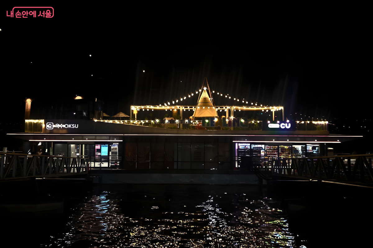 캄캄한 밤, 조명을 환하게 밝힌 옥수선착장의 야경이다. 루프탑을 장식한 알전구들이 삼각형 모양으로 빛나고 있으며, 선착장 건물의 조명이 강물 위에 반사되어 일렁인다. 왼쪽 상단에는 '옥수 OKSU'라는 간판과 오른쪽에 위치한 편의점의 간판도 밝게 빛나고 있다.