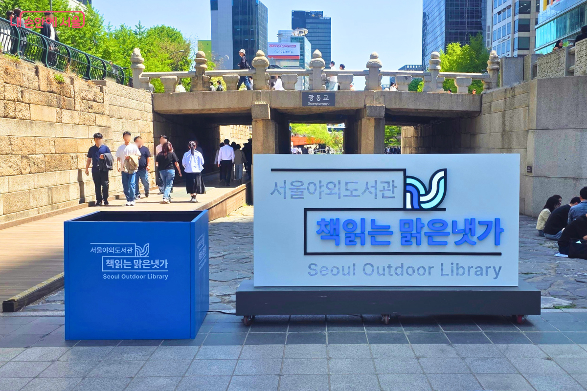 '서울야외도서관 책읽는 맑은냇가'라고 적힌 흰색 대형 간판과 그 옆에 파란색 사각형 입체 간판이 나란히 놓여 있다. 간판 뒤쪽으로는 '광통교'라는 현판이 붙은 석조 다리가 보이며, 다리 위를 건너가는 시민들의 모습이 촬영되었다.