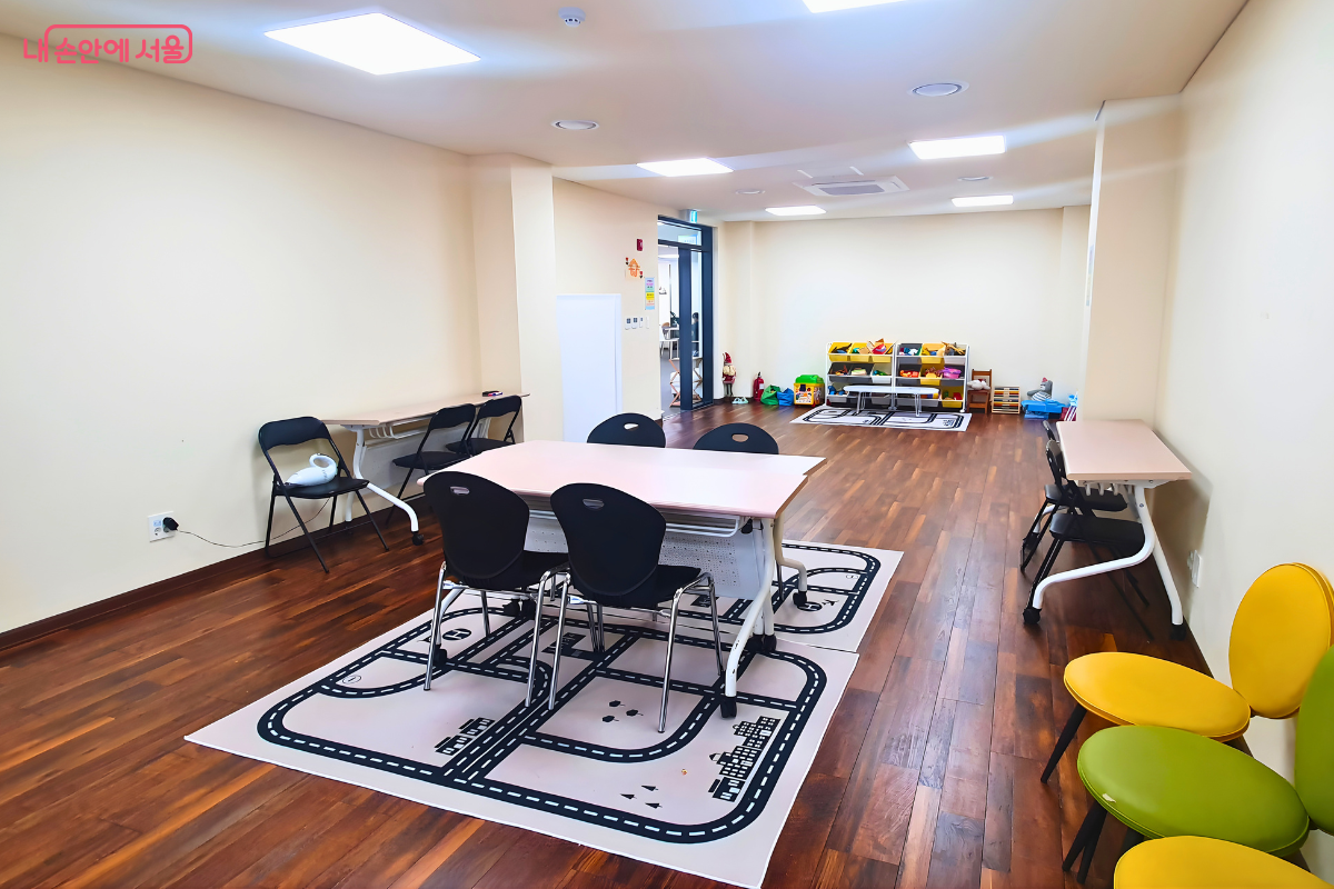 2층 마을 카페 내부와 연결된 다목적실의 모습이다. 나무 재질의 바닥 위에는 아이들이 자동차 놀이를 할 수 있는 도로 모양의 대형 카펫이 깔려 있고, 그 주위로 회의나 수업을 할 수 있는 테이블과 의자들이 놓여 있다. 방 한쪽 구석에는 아이들을 위한 장난감 정리함과 알록달록한 색상의 의자들이 배치되어 있어, 주민들의 소모임이나 어린이 교육 프로그램 장소로 활용됨을 보여준다.