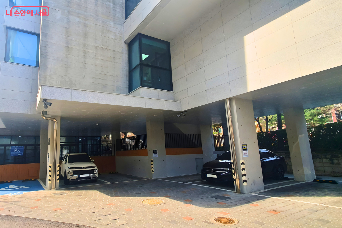 도서관 건물 1층 하단에 위치한 필로티 주차장 전경이다. 기둥 사이로 주차 구역이 구획되어 있으며, 왼쪽 바닥에는 파란색 바탕에 휠체어 문양이 그려진 장애인 전용 주차 구역이 선명하게 보인다. 주차 공간은 장애인 전용 1대를 포함하여 총 7대만 주차가 가능한 규모임을 보여주며, 주변 벽면에는 방문객들의 주차 협조를 요청하는 안내문들이 부착되어 있다.