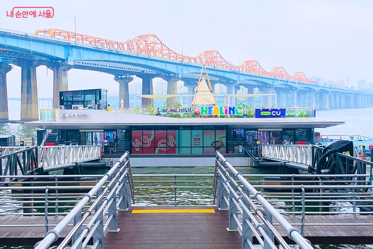 한강 위에 떠 있는 하얀색 옥수선착장 건물의 낮 전경이다. 나무 데크로 된 진입로 정면으로 선착장이 보이며, 건물 위 루프탑에는 'SEOUL SPRING FESTIVAL', 'HEALING'이라고 적힌 알록달록한 간판들이 세워져 있다. 선착장 뒤편으로는 주황색 트러스 구조의 동호대교가 길게 뻗어 있다.