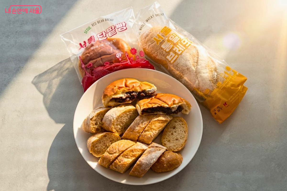 환한 햇살이 비치는 창가 쪽 테이블 위에 흰색 접시가 놓여 있고, 그 안에 서울빵 2종이 시식을 위해 손질된 상태로 담겨 있다. 접시 위에는 먹기 좋게 슬라이스된 통밀브레드와 속이 꽉 찬 단팥 앙금이 보이는 단팥빵 반쪽이 가지런히 놓여 있다. 접시 바로 뒤편에는 내용물을 꺼낸 '서울 단팥빵'과 '서울 통밀브레드'의 빈 패키지가 각각 배치되어 있어 어떤 제품을 시식하는지 명확히 보여준다.