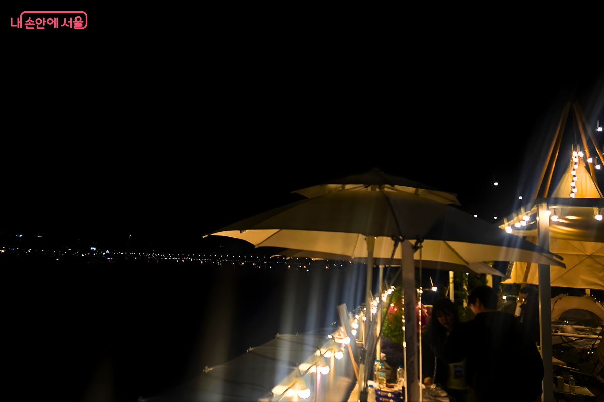 밤이 된 루프탑에서 시민들이 휴식을 취하는 모습이다. 커다란 하얀색 파라솔이 펼쳐져 있고, 그 아래로 조명을 받은 시민들의 실루엣이 보인다. 오른쪽에는 조명이 켜진 원뿔형 텐트 구조물이 보이며, 강 건너편 도심의 불빛들이 멀리 수평선처럼 펼쳐져 있다.