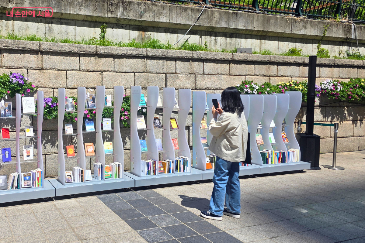 한 시민이 휴대폰을 들어 흰색 물결 모양 서가에 진열된 책들을 촬영하고 있다. 서가 위쪽 돌벽에는 보라색과 분홍색 꽃들이 피어 있다.