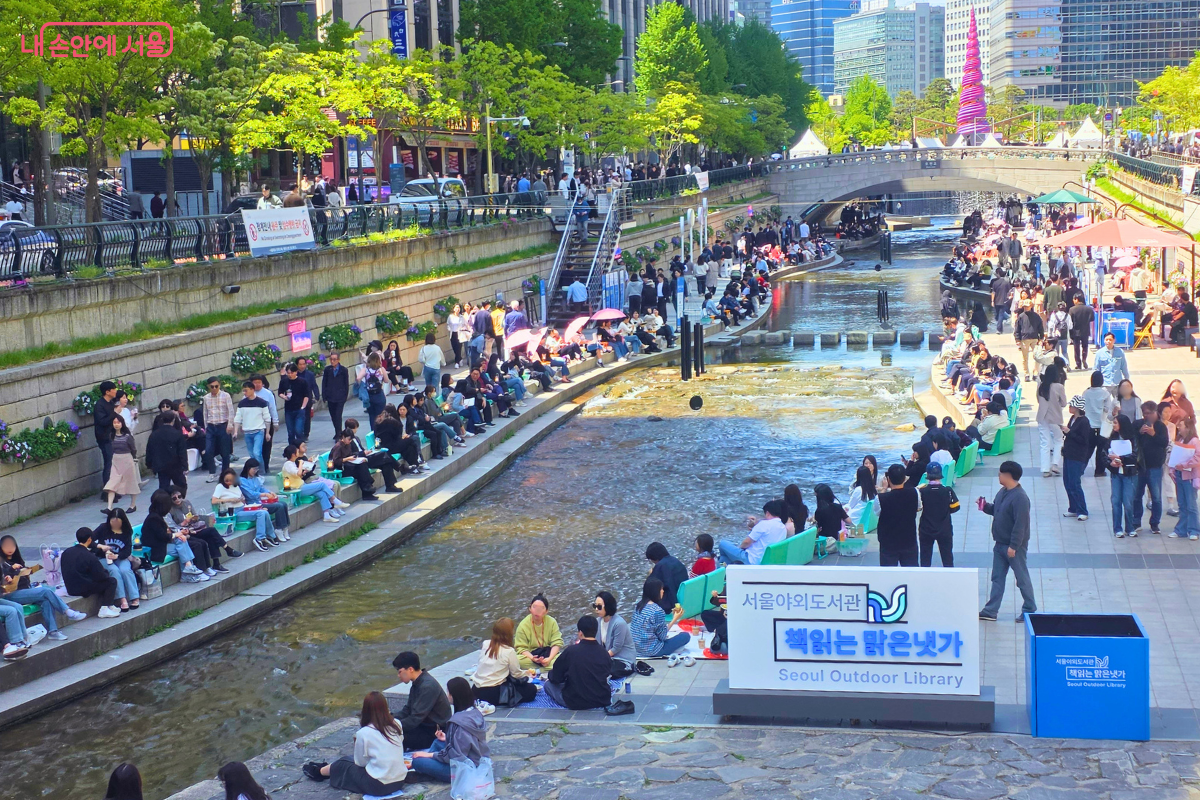 광통교 위에서 하천 상류 방향을 내려다본 풍경이다. 하천 양옆 산책로를 따라 민트색 의자들이 줄지어 배치되어 있고, 시민들이 자리를 잡고 앉아 있다. 우측 하단에는 '서울야외도서관 책읽는 맑은냇가'라고 적힌 흰색 사각형 입간판이 세워져 있으며, 멀리 배경에는 도심 빌딩들과 청계광장의 나선형 조형물이 보인다.