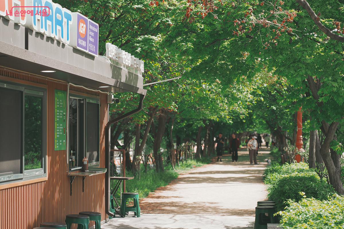 울창한 나무들이 터널을 이룬 흙길 산책로 옆으로 간판이 달린 작은 매점 건물이 있는 평화로운 풍경 &copy;김성무